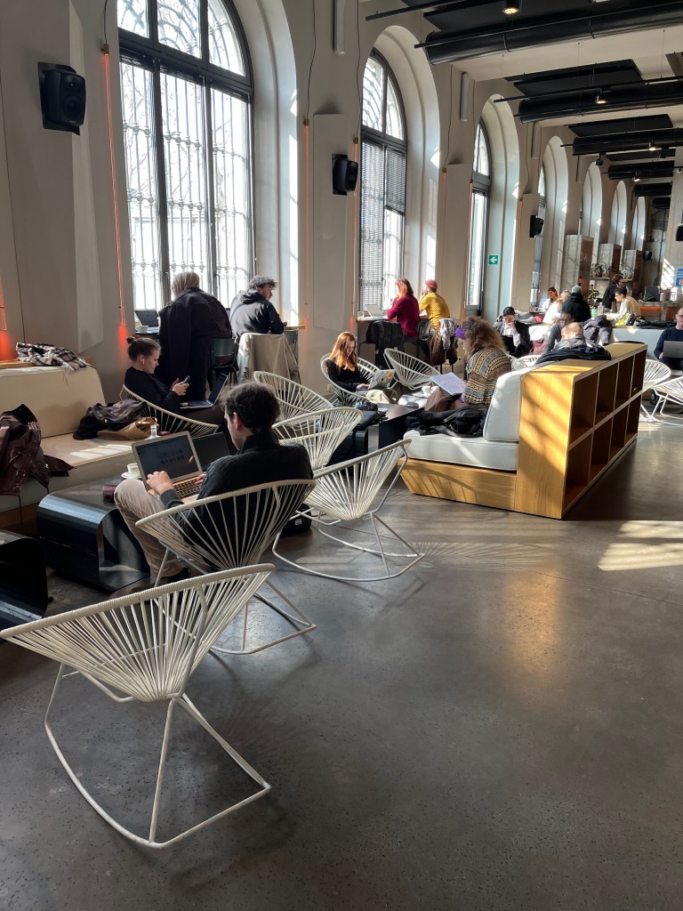 Large room lit by sunlight coming through tall and wide windows. People sitting at high counters, on butterfly chairs and on lounge chairs, working at their laptops.