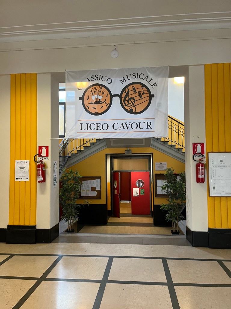 Indoor entrance hallway of Liceo Classico Musicale Cavour (banner) in Turin, Italy. The banner is hanging from the top of the doorway leading to a double staircase in the background and the red doors with porthole windows - left door open - to the auditorium. The support of the doorway sports a fire extinguisher on each side. 